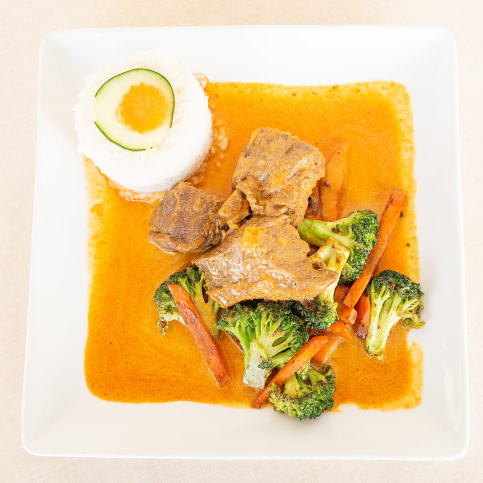 Sliced beef with broccoli and carrots in orange curry sauce, served alongside molded white rice topped with cucumber and orange.
