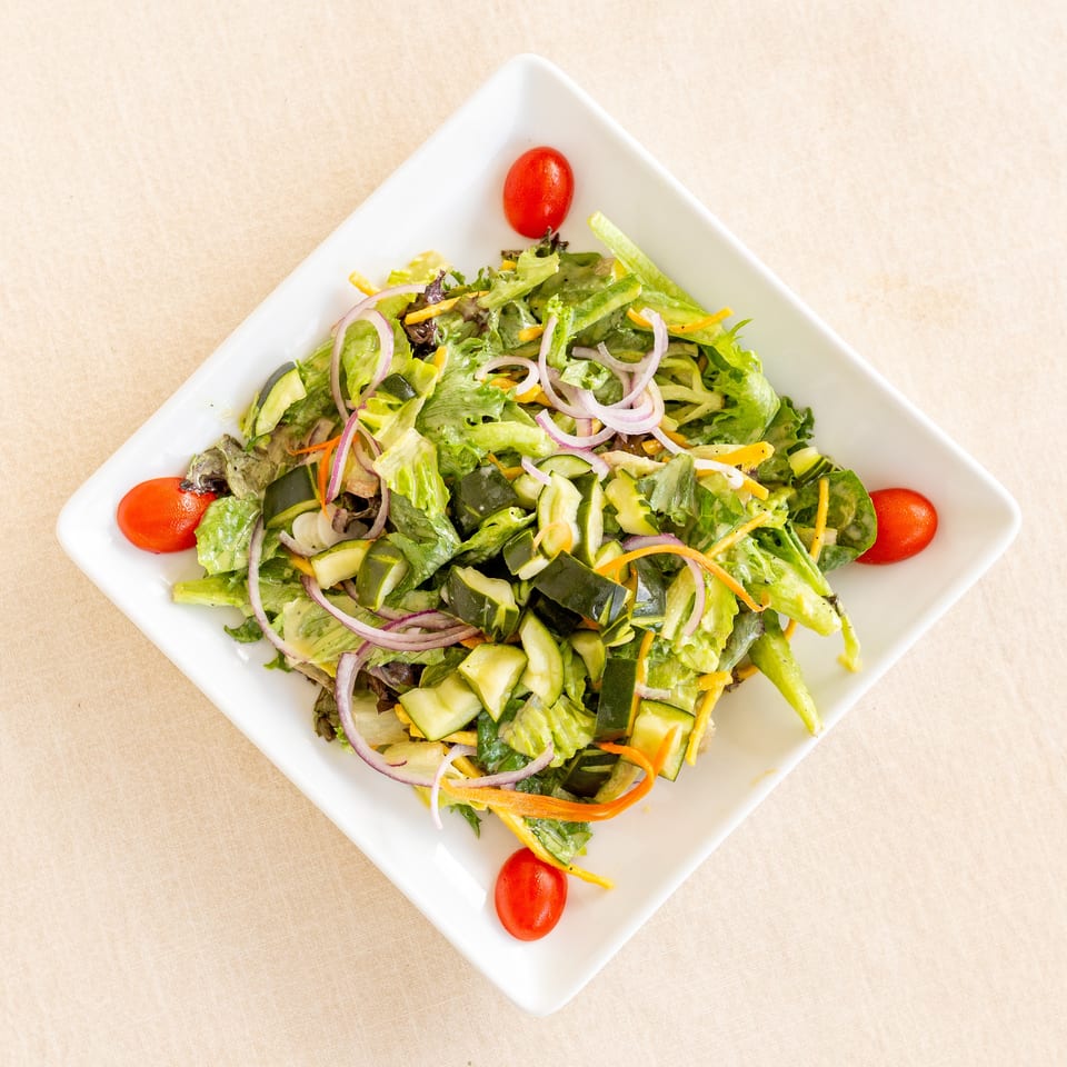Fresh garden salad with lettuce, cucumber, red onion, carrots, zucchini, and cherry tomatoes on a white square plate.