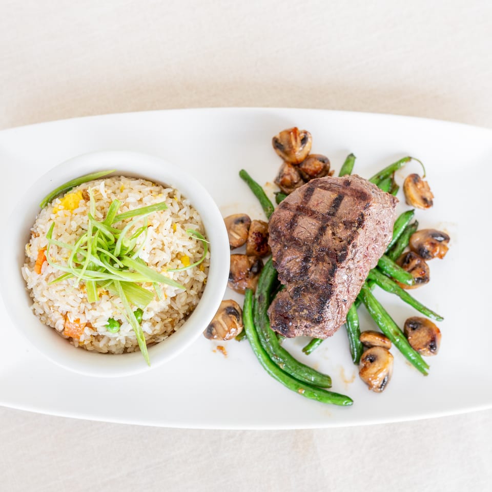 Grilled steak served with fried rice, green beans, and sautéed mushrooms on a white rectangular plate.
