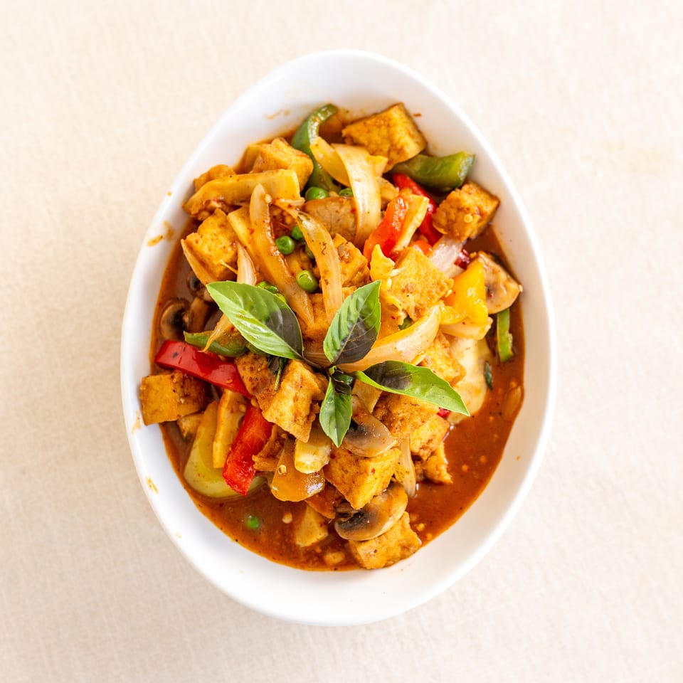 Thai-style red curry with tofu, bell peppers, onions, mushrooms, peas, and fresh basil in a white bowl.