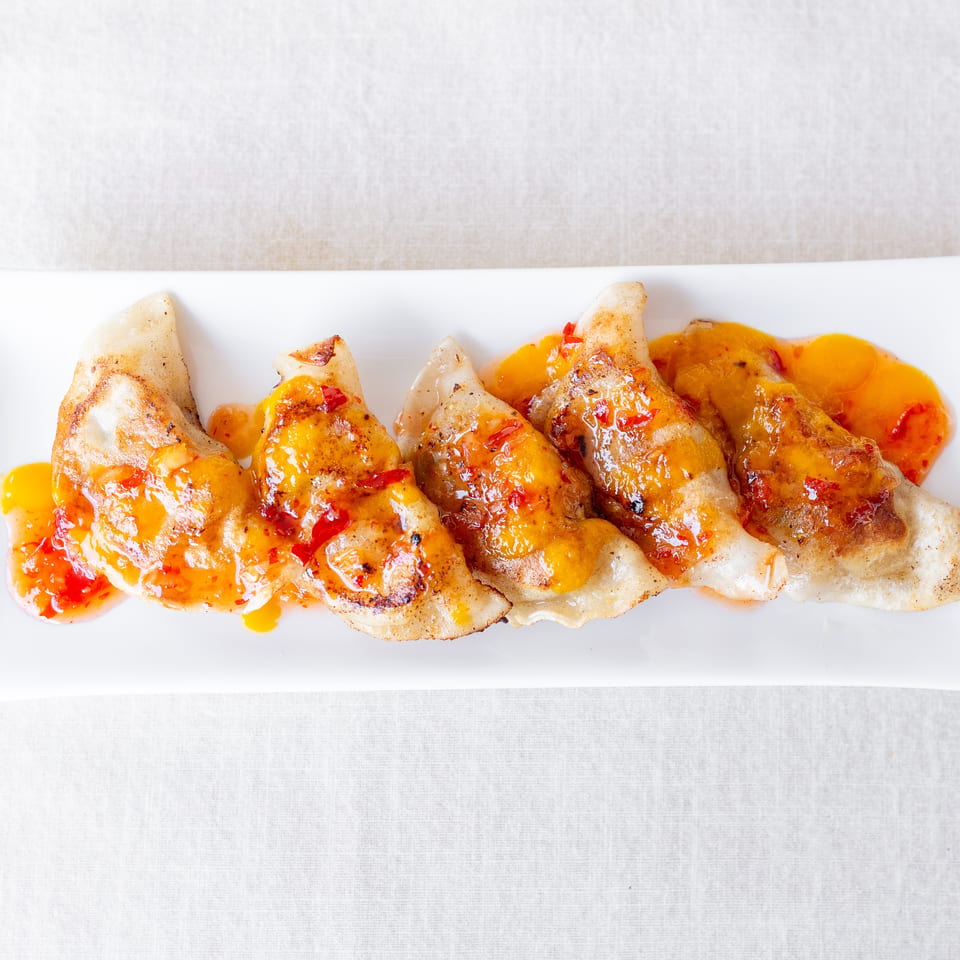 Pan-fried dumplings drizzled with glossy orange chili sauce and red pepper pieces on a white rectangular plate.