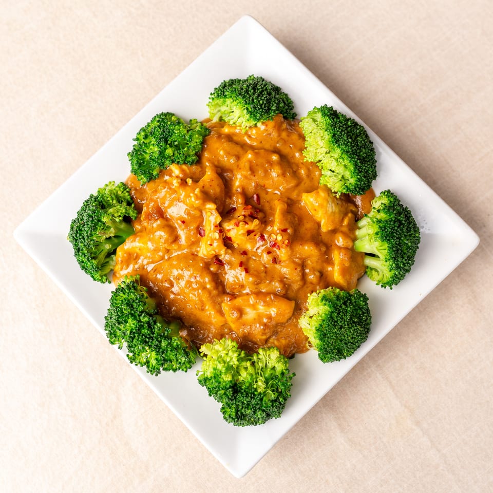 Creamy orange-sauced chicken topped with red pepper flakes and surrounded by steamed broccoli on a white square plate.