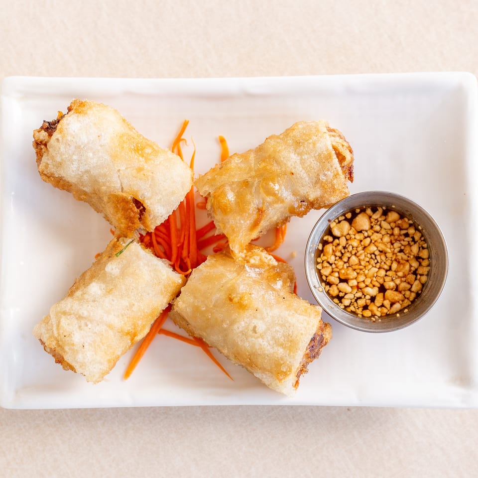 Crispy fried spring rolls arranged over shredded carrots with a bowl of peanut dipping sauce on a white plate.